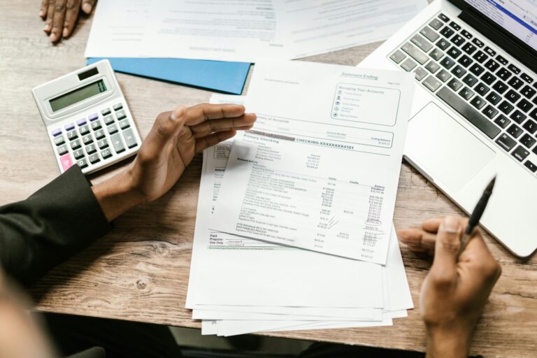 Person reviewing financial documents with calculator and laptop on wooden desk