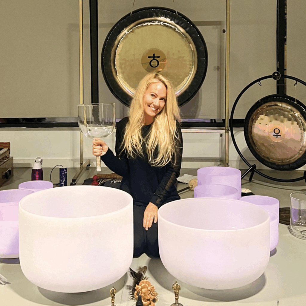 Photo of yoga, meditation, and sound healing educator Jenn Vagios seated with large bowls and holding a large glass.
