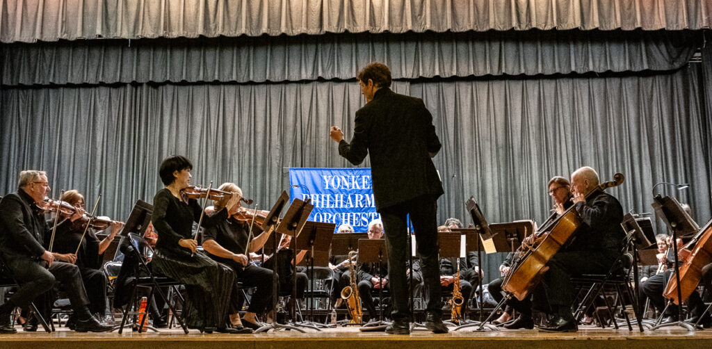 Image of an orchestra on stage, a sign behind them reads "Yonkers Philharmonic Orchestra" 