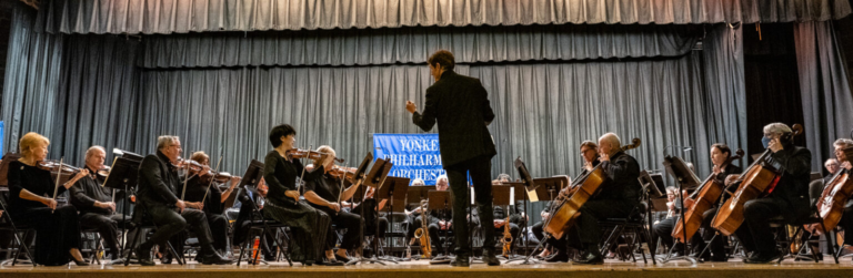 Image of an orchestra on stage, a sign behind them reads "Yonkers Philharmonic Orchestra"